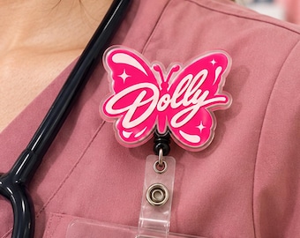 May include: A butterfly-shaped badge reel featuring a vibrant pink acrylic design with white accents and the word Dolly written in a cursive script across the center. The badge reel includes a clear plastic strap with a metal snap button attachment.