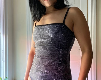 May include: A black and white swimsuit with a geometric pattern. The swimsuit has thin straps and a square neckline.