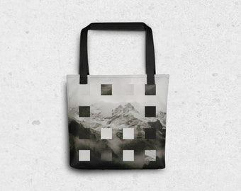 May include: A white tote bag with black straps features a pixelated image of a mountain range with snow-capped peaks and fog. The image is made up of black, white, and grey squares.