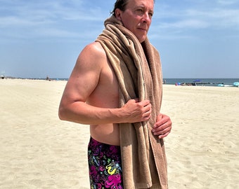 May include: A man on a beach wearing a pair of black swimming trunks with a colourful octopus print and a brown towel draped over his shoulders.