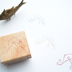May include: A wooden stamp with a carved design of an octopus with tentacles. The stamp is on a white surface with three other octopus designs in different colors.