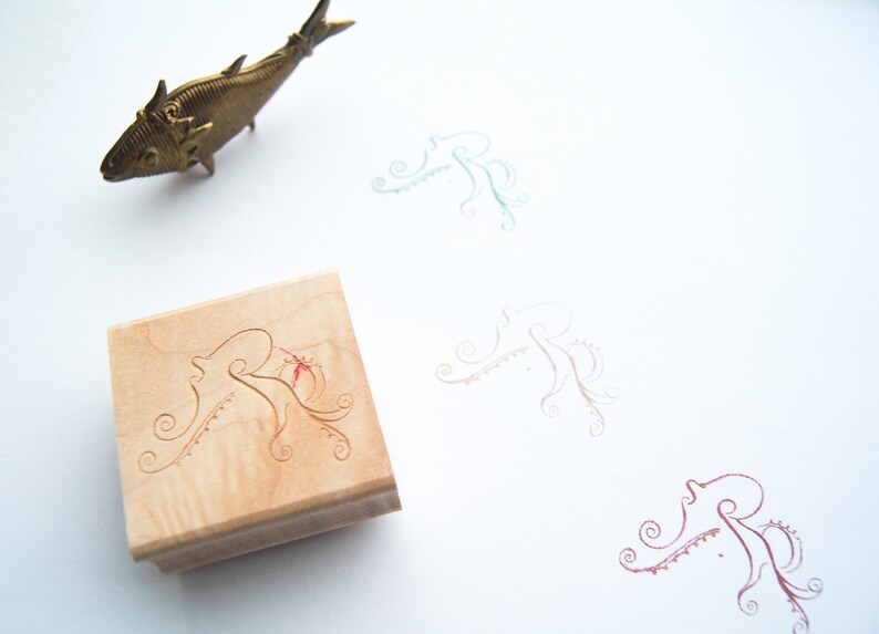 May include: A wooden stamp with a carved design of an octopus with tentacles. The stamp is on a white surface with three other octopus designs in different colors.