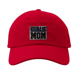 May include: A bright red baseball cap featuring a rectangular patch on the front center. The patch has a grey and black camouflage pattern background with the text GOALIE MOM embroidered in bold, dark navy blue capital letters across the center.