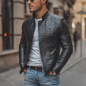 Puede incluir: Una chaqueta negra con un patrón texturizado de piel de cocodrilo, que presenta un cuello mao, mangas largas y un cierre frontal de botones. La chaqueta se lleva sobre una camiseta blanca lisa y unos vaqueros azul claro con un cinturón de cuero marrón.