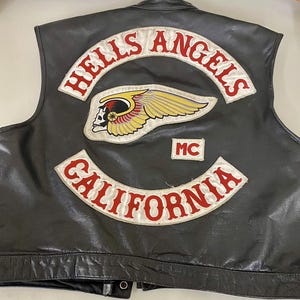 May include: A black leather vest featuring embroidered patches on the back. The top patch reads HELLS ANGELS in red text on a white background. Below is a winged skull emblem, a small MC patch, and a bottom patch reading CALIFORNIA in red.