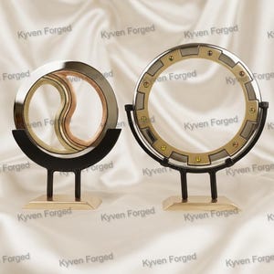 May include: Two decorative metal sculptures on stands. The left sculpture features a silver and gold yin-yang style circular design. The right sculpture is a larger silver and gold gear-shaped ring with small gold studs, both mounted on black and gold bases.