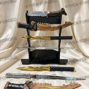 May include: A black display stand holding three knives, with three additional knives placed in front. The collection includes a serrated blade with a horn handle, two bone-textured knives with black wraps, two gold-toned spiral knives, and two silver-toned blades.