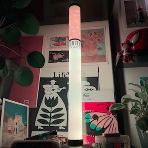 Könnte beinhalten: Eine hohe, zylindrische, beleuchtete Stehlampe mit weißem Sockel und einem oberen Abschnitt mit rosa Muster. Die Lampe zeigt den Text "OUR TEX LIGHTS" in schwarzer Schrift auf ihrem weißen Mittelteil und steht vertikal als Lichtquelle.