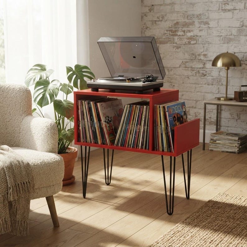 May include: A red wooden record player stand with black metal hairpin legs, featuring two open storage compartments filled with vinyl records and a turntable with a clear dust cover placed on the top surface of the unit.