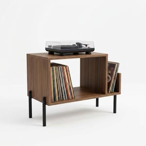 May include: A walnut-finished wooden record player stand with four black metal legs. The unit features a top surface holding a black turntable with a clear dust cover, a large central storage compartment for vinyl records, and a side slot.