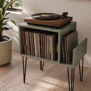 May include: A sage green wooden record player stand with black metal hairpin legs. The stand features a large rectangular storage compartment filled with vinyl records and a smaller side slot for additional albums, topped with a walnut turntable.