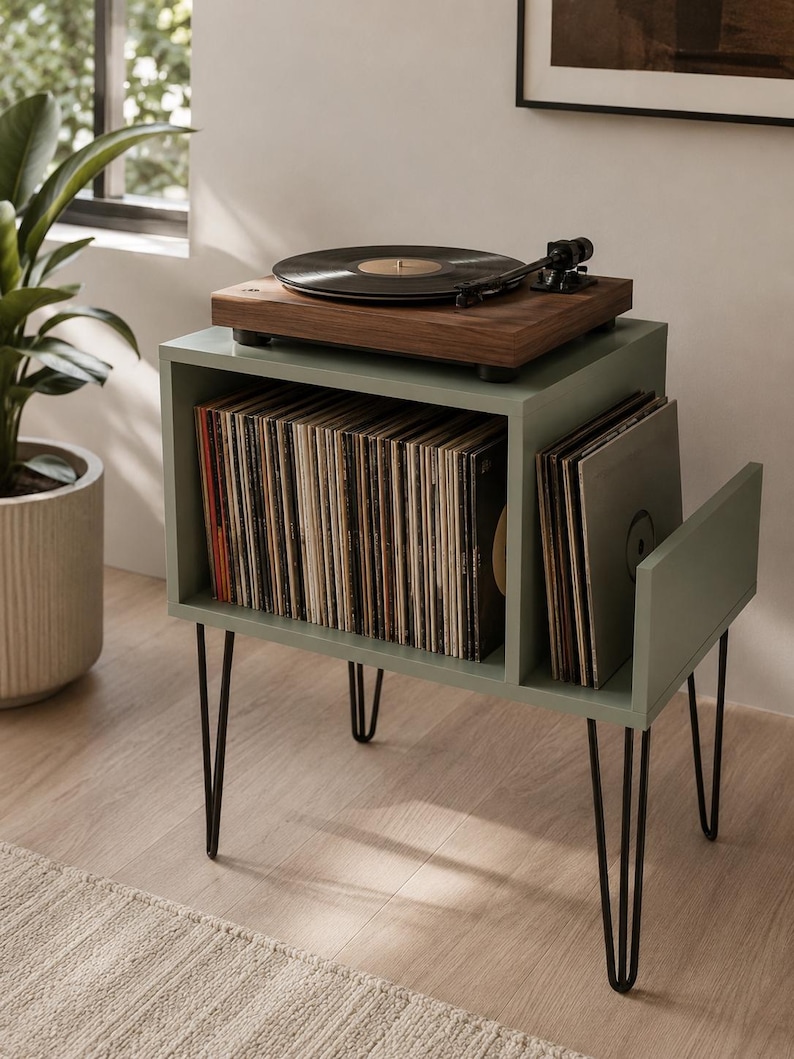 May include: A sage green wooden record player stand with black metal hairpin legs. The stand features a large rectangular storage compartment filled with vinyl records and a smaller side slot for additional albums, topped with a walnut turntable.