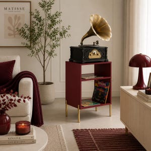 May include: A dark red side table with two open shelves and four gold-toned legs. The table features a black vintage-style gramophone with a large brass horn on top. The shelves contain books and a colourful record sleeve, providing storage space.
