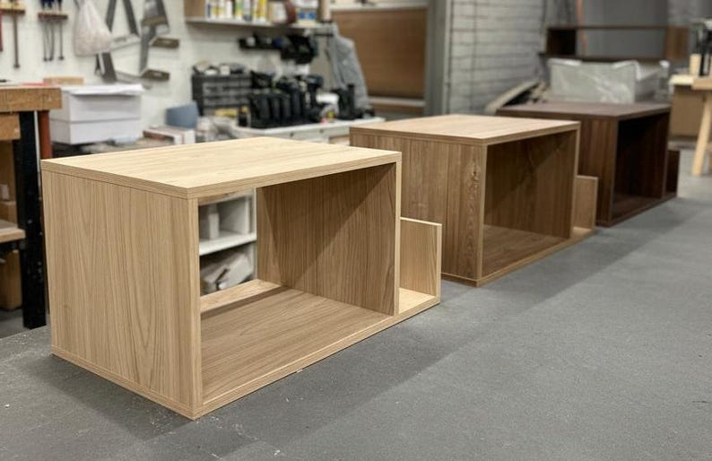 May include: Three open-faced wooden storage units with a rectangular design. Each unit features a light brown wood grain finish and a side compartment. The units are arranged in a row, showcasing a hollow interior space for storage.