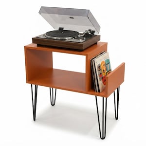 May include: A wooden record player with a clear dust cover sits atop an orange side table. The table features a rectangular open storage compartment, a side slot holding several vinyl records, and four black metal hairpin legs.