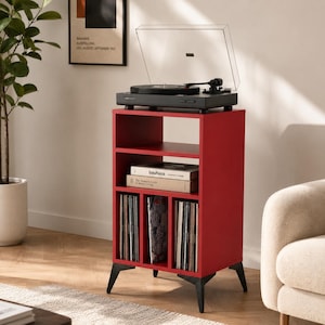 May include: A red wooden record player stand with black angled legs, featuring an open top shelf, a middle shelf holding two books titled Bauhaus and The Monocle Guide to Better Living, and three vertical compartments filled with vinyl records.