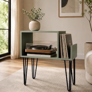 May include: A sage green wooden record player stand with a rectangular open shelf and a vertical side compartment for vinyl records. The unit features four black metal hairpin legs and holds a turntable, a book, and a small ceramic vase.