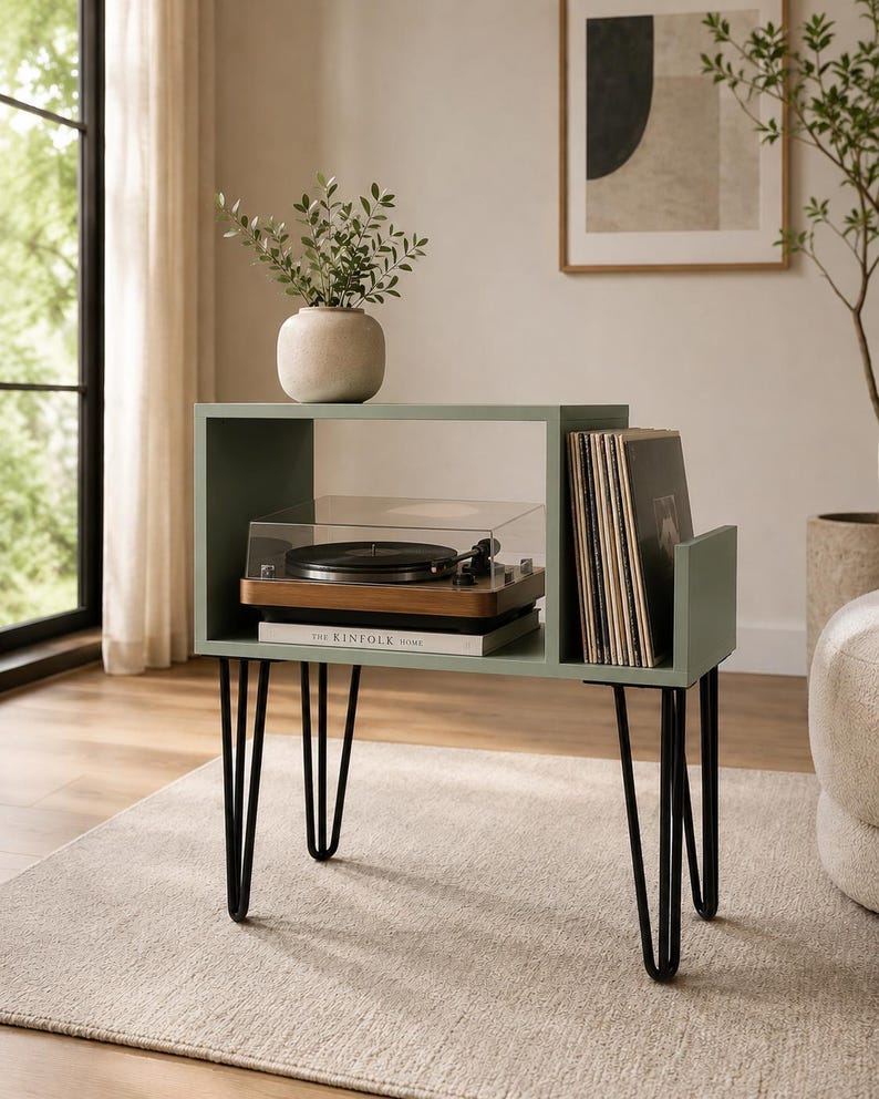 May include: A sage green wooden record player stand with a rectangular open shelf and a vertical side compartment for vinyl records. The unit features four black metal hairpin legs and holds a turntable, a book, and a small ceramic vase.