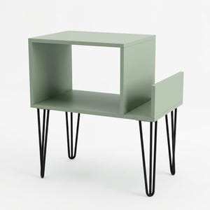 May include: A sage green side table featuring a rectangular open storage compartment and an attached lower side shelf. The unit is supported by four thin, black metal hairpin legs that provide a stable base for the wooden structure.
