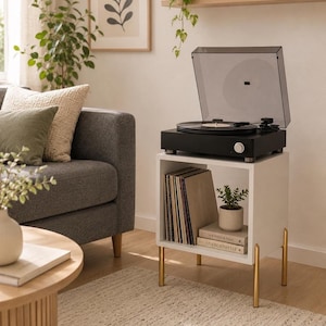 May include: A white square side table with four gold-toned metal legs, featuring an open storage compartment containing a collection of vinyl records, two stacked books, and a small potted plant, topped with a black turntable with a clear lid.