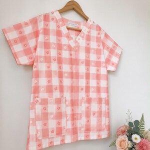 May include: A short-sleeved scrub top featuring a pink and white gingham check pattern with small white flower accents. The garment has a V-neckline and two large square patch pockets on the front, hanging on a wooden hanger.
