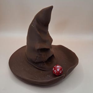 May include: A brown, pointed, slouchy hat with a wide brim and a textured, fabric-like appearance. A red, twenty-sided die with white numbers is positioned on the brim of the hat. The hat features a deep fold and a prominent, curved peak.