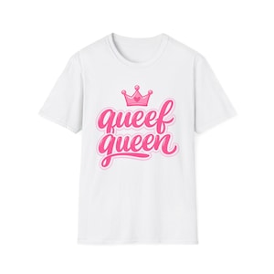 May include: A white short-sleeved t-shirt featuring a pink graphic print on the chest. The design includes a small pink crown with a heart detail above the stylized pink text that reads "queef queen" in a cursive, bold, and rounded font style.