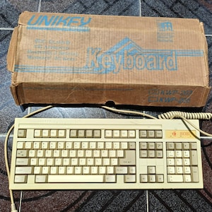 May include: A beige computer keyboard with a coiled cable and a matching cardboard box. The box features the text UNIKEY, Keyboard, and KWP-200. The keyboard has a standard QWERTY layout with a numeric keypad and a small orange logo on the right side.