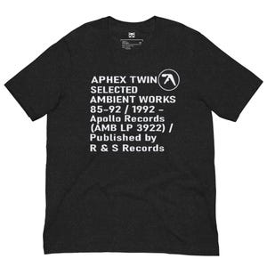 May include: A charcoal grey short-sleeve t-shirt featuring white text that reads APHEX TWIN SELECTED AMBIENT WORKS 85-92 / 1992 - Apollo Records (AMB LP 3922) / Published by R & S Records, along with the Aphex Twin logo in a white circle.