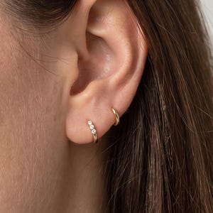 May include: A pair of gold hoop earrings worn in the earlobe. One hoop is plain gold, while the other features three small, clear, round-cut stones set in a row along the front of the hoop. Both earrings have a polished, metallic finish.