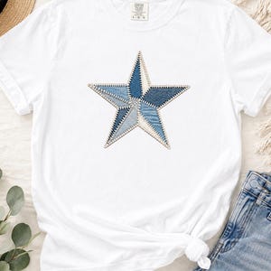 Puede incluir: Una camiseta blanca de manga corta con un gráfico central de estrella compuesto por varios parches de tela en tonos azules que recuerdan al denim. La estrella está delineada con una costura blanca visible, creando un aspecto texturizado.
