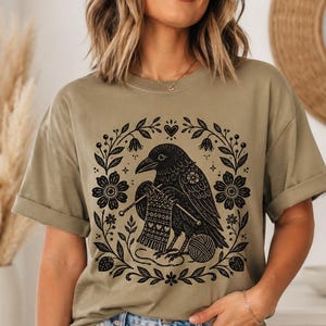 May include: An olive green short-sleeve t-shirt featuring a black graphic print of a raven knitting with needles and yarn. The bird is centered within a circular frame composed of botanical elements, including flowers, leaves, and small hearts.
