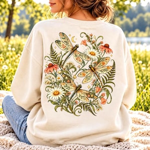 May include: A cream-colored crewneck sweatshirt featuring a large back graphic of three dragonflies surrounded by various wildflowers, ferns, and small stars. The design includes orange, yellow, and white floral elements with green foliage.