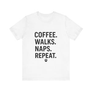 Könnte beinhalten: Ein weißes kurzärmeliges T-Shirt mit schwarzem Text, der mittig auf der Brust steht: COFFEE. WALKS. NAPS. REPEAT. Darunter befindet sich ein kleines schwarzes Pfotenabdruck-Symbol. Das T-Shirt hat einen Rundhalsausschnitt und eine Standardpassform.