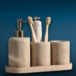 Peut inclure: Un ensemble d'accessoires de salle de bain comprenant un distributeur de savon en pierre beige texturée, un porte-brosse à dents assorti avec deux brosses en bambou et un tube blanc, et un gobelet, le tout sur un plateau rectangulaire.