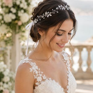 May include: A silver-toned hair vine featuring delicate metal leaves and small clear crystals, arranged in a flexible vine pattern. The accessory is secured with a thin white ribbon tied at the back, resting atop dark hair styled in an updo.