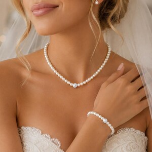 May include: A necklace and bracelet set featuring uniform white pearls and a single central spherical charm encrusted with clear crystals. The necklace rests at the base of the neck, and the matching bracelet is worn on the wrist.