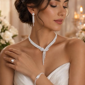 May include: A silver-toned jewellery set featuring a necklace with a central floral motif and cascading crystal fringe, matching drop earrings with a marquise-cut stone, a delicate crystal bracelet, and a ring with a marquise-cut centre stone.