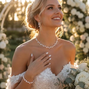 May include: A jewellery set consisting of a necklace, a bracelet, and drop earrings, all featuring small white pearls arranged in a delicate, repeating pattern on a thin metallic chain. The pieces are worn with a white lace off-the-shoulder dress.