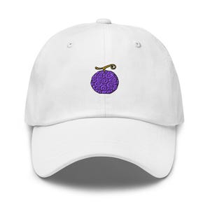 Könnte beinhalten: Eine weiße Baseballkappe mit einer kleinen, mittig platzierten Stickerei einer violetten Frucht mit Wirbelmuster und goldenem Stiel. Die Kappe hat einen gebogenen Schirm, ein strukturiertes Vorderteil und Belüftungsösen an der Krone.