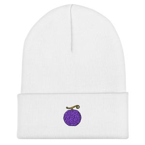 May include: A white beanie featuring a small, centered embroidery of a purple, round fruit with a swirling texture and a brown stem. The knit cap has a folded cuff at the bottom, providing a clean, simple look with the detailed graphic.