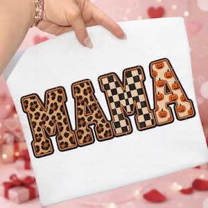 May include: A white t-shirt featuring the word MAMA in large block letters. The first two letters have a brown and black leopard print, the third letter has a black and white checkerboard pattern, and the final letter has an orange pumpkin print.