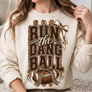 Op de afbeelding: Een crèmekleurige sweater met ronde hals en een grafische print met de tekst "RUN the DANG BALL" in bruine blokletters. Het ontwerp bevat een bruine football met een luipaardprint strik en een grote gouden strik, geplaatst tegen luipaardprint strepen.