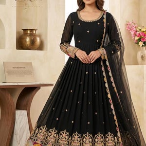 May include: A black floor-length Anarkali dress featuring long sleeves with intricate gold, pink, and yellow embroidery at the cuffs and hem. The bodice has a round neckline with gold detailing, and the skirt includes a matching sheer black dupatta.
