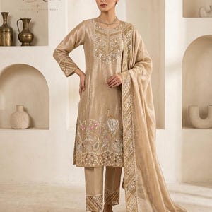 May include: A beige three-piece ensemble featuring a long-sleeved tunic with intricate gold embroidery, a round neckline, and a matching straight-leg trouser. The outfit includes a sheer, embroidered dupatta draped over the shoulder with detailed borders.