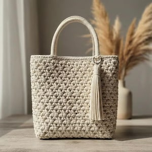 May include: A cream-colored, hand-crocheted tote bag featuring a thick, textured stitch pattern, two sturdy rounded handles, and a matching long decorative tassel attached to a silver-toned metal ring on the front of the bag.