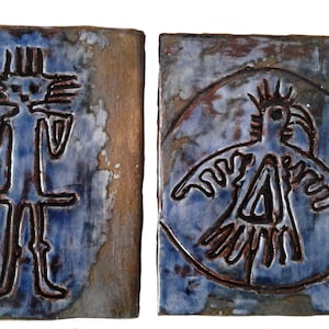 May include: Two ceramic tiles with blue and brown glaze. The tile on the left has a stylized human figure drawn in brown. The tile on the right has a stylized bird figure drawn in brown inside a circle.