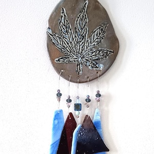 May include: A decorative wind chime featuring a brown ceramic top with a detailed leaf design. Below, glass shapes in blue, burgundy, and dark hues hang with beaded accents. A green, leaf-shaped charm dangles at the bottom.