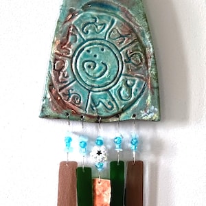 May include: A handcrafted wind chime with a turquoise ceramic top featuring an embossed design. Below hang rectangular pieces of brown, green, and copper-colored materials, accented with blue and white beads. A decorative item.