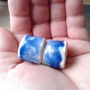 May include: Two cylindrical ceramic beads featuring a glossy white base with irregular, mottled blue watercolor-style patterns. The beads have a smooth, reflective finish and are shaped like small, thick tubes with rounded edges and a glazed surface.